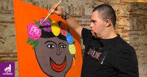 Color photo of Mexican artist Alan Tellez painting a colorful picture of a face with a big smile and flowers in her hair. 