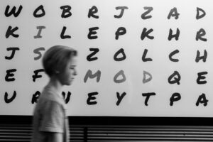 Black and white photo of a young teen walking past a large board with the letters of the alphabet displayed there.