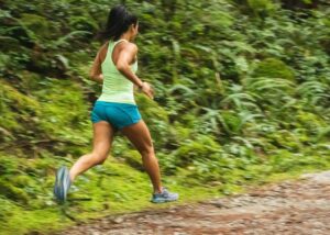 Color photo of a young woman jogging on a dirt path next to green bushes and trees. 